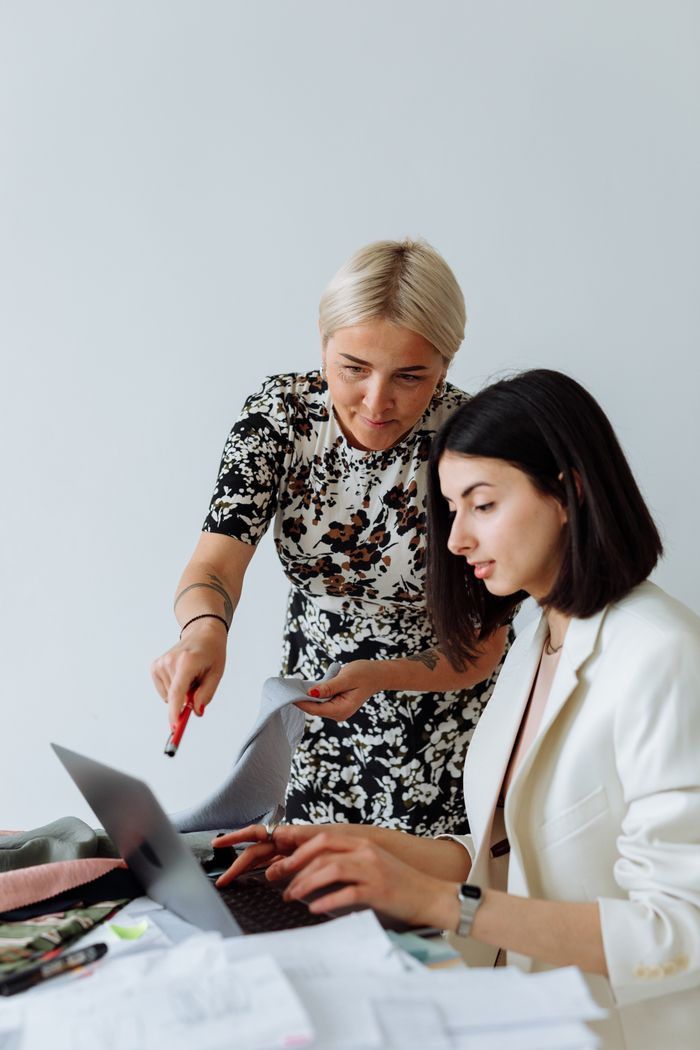 About Two women discussing a project at a desk with a laptop in a modern office setting.